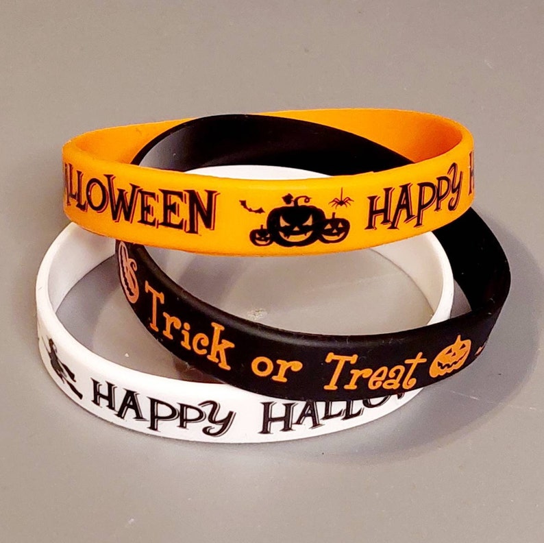 May include: Three Halloween themed silicone wristbands. One orange wristband says "Happy Halloween" with a pumpkin and bats. One black wristband says "Trick or Treat" with a pumpkin. One white wristband says "Happy Halloween" with a witch.