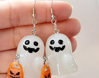 Op de afbeelding: Een paar witte spookoorbellen met zwarte ogen en een glimlachende mond. Elke spook houdt een kleine oranje pompoen vast met een zwart pompoengezicht.