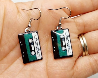 May include: A pair of silver earrings with a green and black cassette tape charm. The charms have the text "Good Vibes" printed on them.
