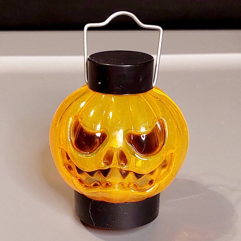 May include: A small orange plastic jack-o'-lantern with a black handle and a scary face. The lantern is lit from inside, creating a warm glow.