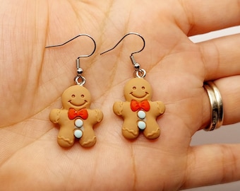 May include: A pair of gingerbread man earrings with red bows and white buttons. The earrings are made of polymer clay and have silver hooks.