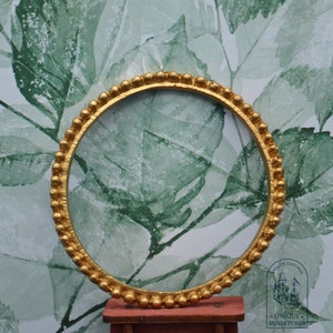 May include: A gold-colored round frame with a beaded edge. The frame is against a green and white floral background.