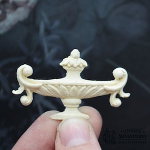 Könnte beinhalten: Eine weiße Keramikurne mit einem dekorativen Deckel und geschwungenen Griffen. Die Urne ist eine Miniatur-Nachbildung einer traditionellen Gartenurne.