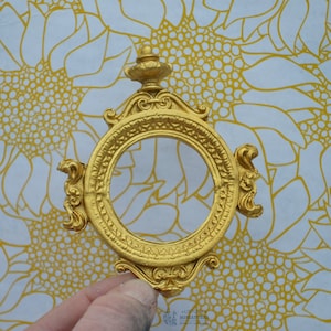 May include: A gold-colored ornate frame with a round opening. The frame has a decorative design with a finial on top.