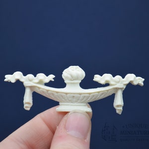 May include: White resin decorative trim with a central floral design and two scrolled side pieces. The trim is designed for use in dollhouse or miniature projects.