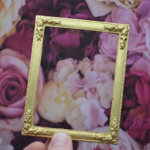 以下が含まれることがあります： 装飾的なディテールが施された金色の写真フレーム。フレームはピンクと白の花の背景に置かれています。
