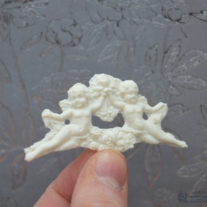 May include: White ceramic cherub figurine with wings, holding a heart-shaped object. The figurine is detailed with floral accents and has a cutout in the center.