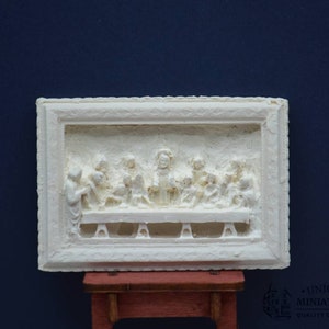 May include: A white plaster relief sculpture depicting the Last Supper. The sculpture is framed in a white plaster frame and is mounted on a wooden easel.