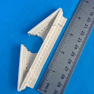 Può includere: Dettaglio architettonico in gesso bianco, che ricorda una cornice o una modanatura, con sculture intricate. Il dettaglio è lungo circa 7,6 cm ed è mostrato accanto a un righello per la scala.