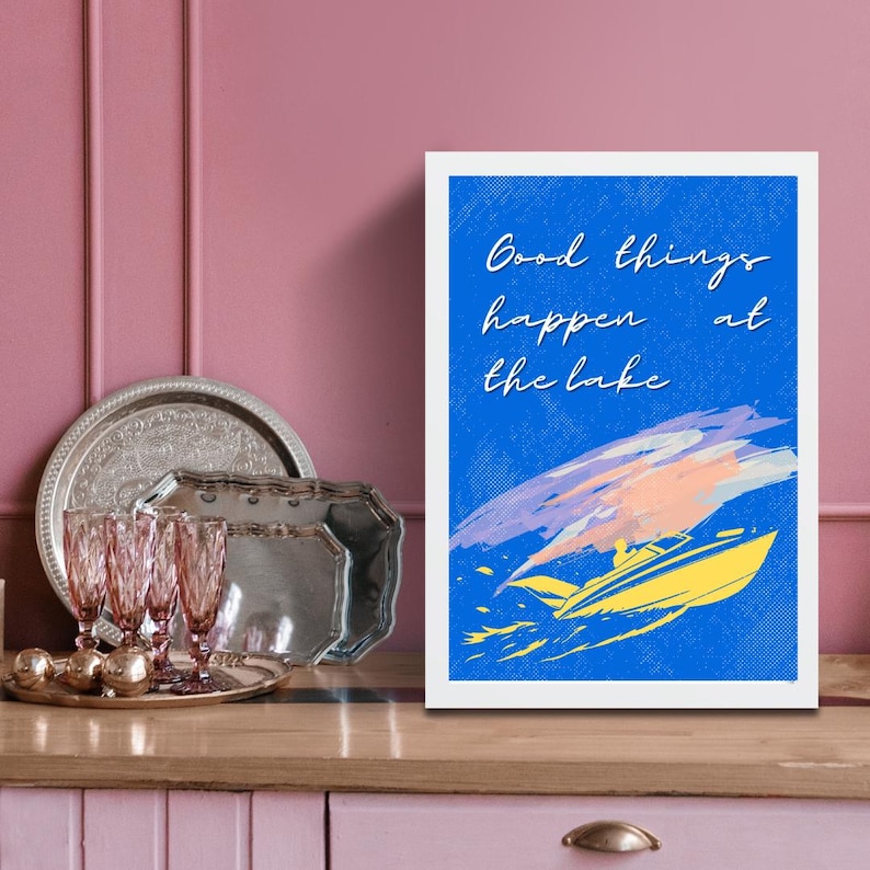 以下が含まれることがあります： 青い背景に「Good things happen at the lake」の文字が入った額入りのアートプリント。 黄色のボートと、紫、ピンク、オレンジ色の抽象的な筆致が特徴です。 背景には銀色のトレイとピンク色のグラスがあります。