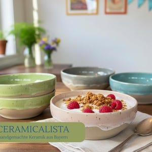 Op de afbeelding: Een verzameling handgemaakte keramische kommen in verschillende kleuren, waaronder groen, grijs en blauw. E&eacute;n kom is gevuld met yoghurt, granola en frambozen. De kommen staan op een houten tafel. De tekst op de afbeelding luidt "CERAMICALISTA handgemachte Keramik aus Bayern."