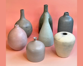 May include: A collection of seven ceramic vases in various shapes and colors. The vases include shades of gray, pink, and green. Some have narrow necks, while others are rounded or cone-shaped. The vases are displayed on a pink background.
