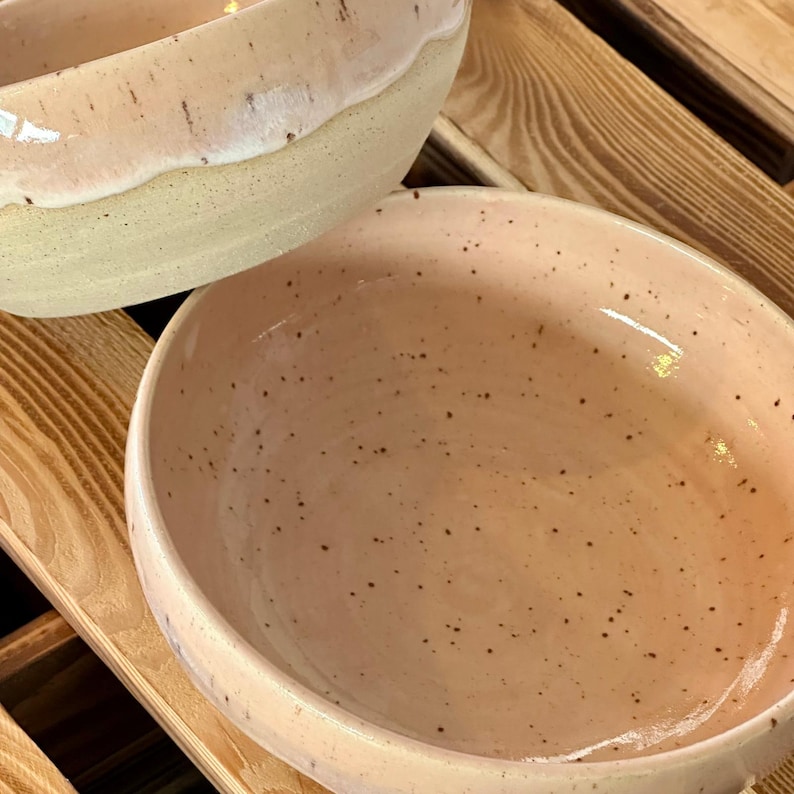 Op de afbeelding: Twee keramische kommen met een gespikkeld, lichtroze interieur en een cr&egrave;mewitte rand. De kommen staan op een houten oppervlak. De kommen hebben een gladde, glanzende afwerking.