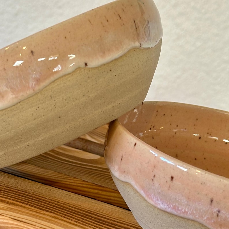 Op de afbeelding: Twee handgemaakte keramische kommen. De kommen hebben een lichtbeige basis met een roze-perzik glazuur op de rand. Het glazuur heeft een glanzende afwerking en een gespikkeld uiterlijk. De kommen staan op een houten oppervlak.