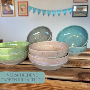 Op de afbeelding: Een verzameling keramische kommen in verschillende kleuren, waaronder groen, grijs, roze en turquoise. De kommen zijn gestapeld op een houten oppervlak. De tekst onderaan luidt "VERSCHIEDENE FARBEN ERH&Auml;LTLICH".