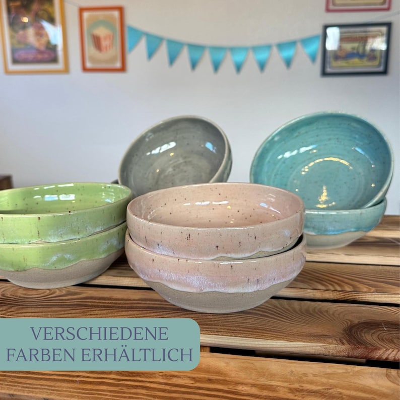 Op de afbeelding: Een verzameling keramische kommen in verschillende kleuren, waaronder groen, grijs, roze en turquoise. De kommen zijn gestapeld op een houten oppervlak. De tekst onderaan luidt "VERSCHIEDENE FARBEN ERH&Auml;LTLICH".