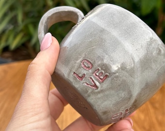 Puede incluir: Una taza de cerámica gris con asa y la palabra "LOVE" escrita en rosa en el lateral.
