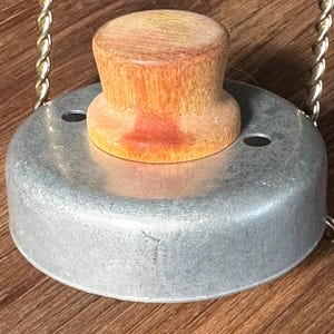 May include: A circular metal biscuit cutter featuring a smooth, light-colored wooden knob handle centered on top. The silver-toned metal base has two small circular holes on opposite sides, and a twisted gold-colored wire frame is attached to the sides.