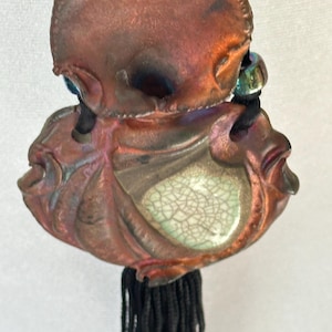 May include: A pendant necklace featuring two interconnected, rounded copper-toned elements with a metallic, iridescent finish. The lower element contains a central, light green crackle-glazed inlay. A black cord and a black tassel are attached.