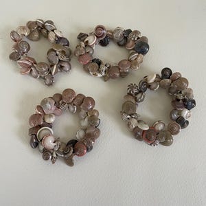 May include: Four decorative seashell napkin rings. Each ring is crafted from a variety of small, naturally colored seashells, including shades of tan, brown, and cream. The shells are arranged in a circular pattern.