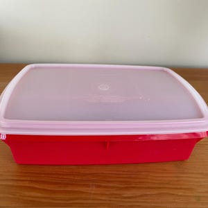May include: A rectangular red food storage container with a clear, slightly translucent lid. The lid has a pink rim and a circular design in the center. The container is sitting on a wooden surface.
