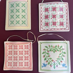 May include: Four square ceramic wall plaques with hanging ribbons. One features a green geometric quilt pattern, one a pink floral grid, one a red geometric pattern, and one a green heart-shaped vine with two blue birds.