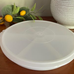 May include: A white plastic lid with a circular design and two side handles. The lid is on a wooden surface, with a branch of lemons and a decorative vase in the background.