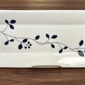 May include: A rectangular white ceramic serving tray featuring a delicate, dark blue botanical vine pattern that stretches horizontally across the center. The design includes small leaves and clusters of berries, presented on a smooth, glossy surface.