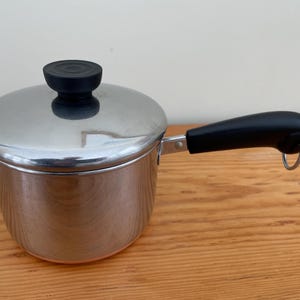 May include: A stainless steel saucepan with a lid and a black handle. The saucepan has a rounded body and a lid with a black knob. The handle is attached to the side of the pan and has a loop at the end. Kitchenware.