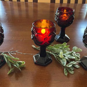 May include: Four ruby red goblets with a textured design, each with a lit candle inside, sit on a wooden surface. The goblets have black stems and bases. Greenery and a twig are arranged between the goblets, creating a decorative display.