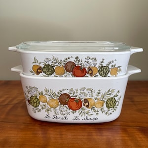 May include: Two white rectangular casserole dishes with a clear glass lid. The dishes are decorated with a colorful vegetable and herb design. The text "Le Persil, Le Sauge" is printed on the front of the bottom dish.