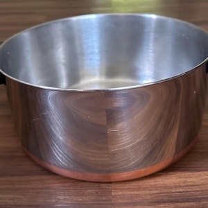 May include: A stainless steel cooking pot with a copper-colored base and black handles. The pot has a shiny, reflective surface and is sitting on a wooden surface. The pot is designed for cooking.