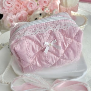 May include: A pink quilted makeup bag with a white zipper and lace trim. A small white bow with a pearl center is on the front of the bag.