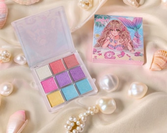 May include: A mermaid-themed makeup set featuring a nine-color eyeshadow palette with pastel shades and a compact with a mermaid illustration. The set is surrounded by pearls and seashells on a satin surface.