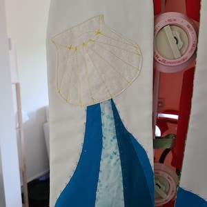 May include: A white fabric panel with a yellow embroidered seashell and blue fabric sewn in a wave pattern below it.
