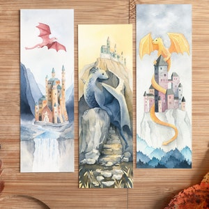 K&ouml;nnte beinhalten: Drei Aquarell-Lesezeichen mit Drachen und Burgen. Das erste Lesezeichen zeigt einen rosa Drachen, der &uuml;ber einer Burg mit einem Wasserfall fliegt. Das zweite Lesezeichen zeigt einen blauen Drachen, der auf einem Felsen mit einer Burg im Hintergrund sitzt. Das dritte Lesezeichen zeigt einen gelben Drachen, der sich um eine Burg auf einem Berg windet.