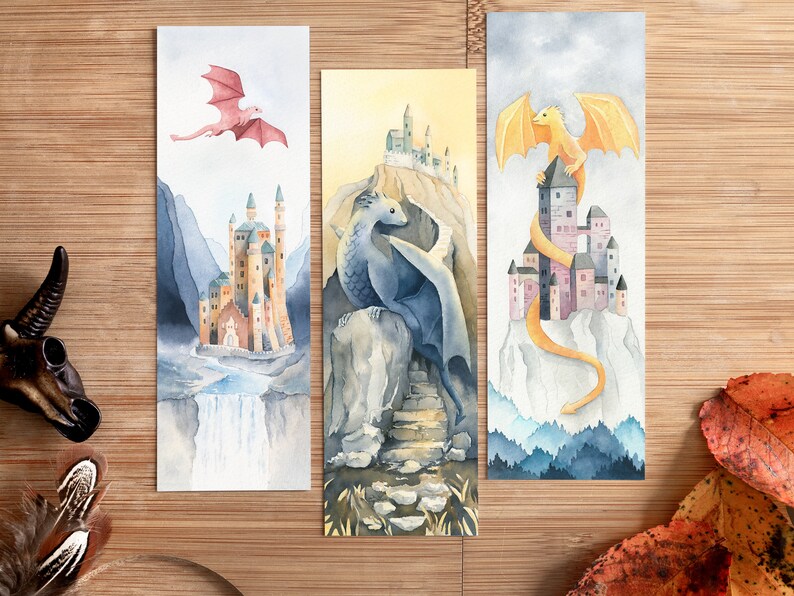 K&ouml;nnte beinhalten: Drei Aquarell-Lesezeichen mit Drachen und Burgen. Das erste Lesezeichen zeigt einen rosa Drachen, der &uuml;ber einer Burg mit einem Wasserfall fliegt. Das zweite Lesezeichen zeigt einen blauen Drachen, der auf einem Felsen mit einer Burg im Hintergrund sitzt. Das dritte Lesezeichen zeigt einen gelben Drachen, der sich um eine Burg auf einem Berg windet.