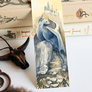 K&ouml;nnte beinhalten: Ein Aquarellbild eines blauen Drachen, der auf einem Felsen sitzt, mit einer Steintreppe, die zu einem Schloss in der Ferne f&uuml;hrt. Der Drache hat Fl&uuml;gel und einen langen Schwanz. Der Hintergrund ist hellgelb.
