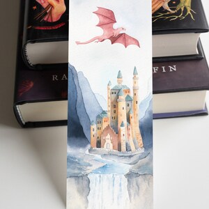 K&ouml;nnte beinhalten: Ein Aquarell-Lesezeichen mit einem rosa Drachen, der &uuml;ber einem Schloss mit einem Wasserfall im Vordergrund fliegt. Das Schloss ist von Bergen umgeben und hat einen hellblauen Himmel im Hintergrund.