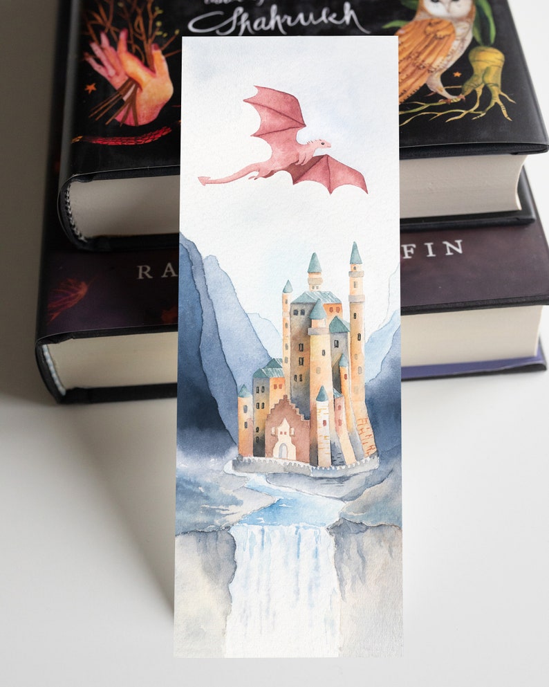 K&ouml;nnte beinhalten: Ein Aquarell-Lesezeichen mit einem rosa Drachen, der &uuml;ber einem Schloss mit einem Wasserfall im Vordergrund fliegt. Das Schloss ist von Bergen umgeben und hat einen hellblauen Himmel im Hintergrund.