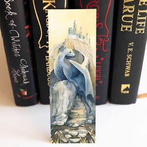 K&ouml;nnte beinhalten: Ein Aquarell-Lesezeichen mit einem blauen Drachen, der auf einem Felsen sitzt, mit einer Steintreppe, die zu einer kleinen Burg auf einer Klippe f&uuml;hrt. Der Hintergrund ist in einem sanften Gelb gehalten.