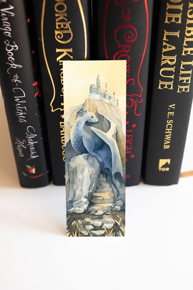 K&ouml;nnte beinhalten: Ein Aquarell-Lesezeichen mit einem blauen Drachen, der auf einem Felsen sitzt, mit einer Steintreppe, die zu einer kleinen Burg auf einer Klippe f&uuml;hrt. Der Hintergrund ist in einem sanften Gelb gehalten.