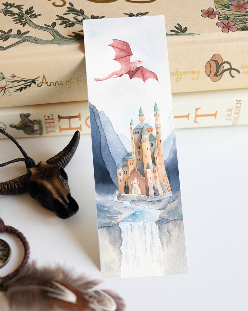 K&ouml;nnte beinhalten: Aquarell-Lesezeichen mit einem rosa Drachen, der &uuml;ber einem Schloss mit einem Wasserfall fliegt. Das Schloss ist von Bergen umgeben und der Himmel ist hellblau.