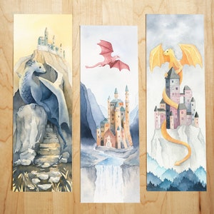 K&ouml;nnte beinhalten: Drei Aquarelle von Drachen und Burgen. Das erste Bild zeigt einen blauen Drachen, der auf einem Felsen sitzt, mit einer Burg im Hintergrund. Das zweite Bild zeigt einen rosa Drachen, der &uuml;ber einer Burg mit einem Wasserfall im Vordergrund fliegt. Das dritte Bild zeigt einen gelben Drachen, der sich um eine Burg mit einem Gebirge im Hintergrund schl&auml;ngelt.