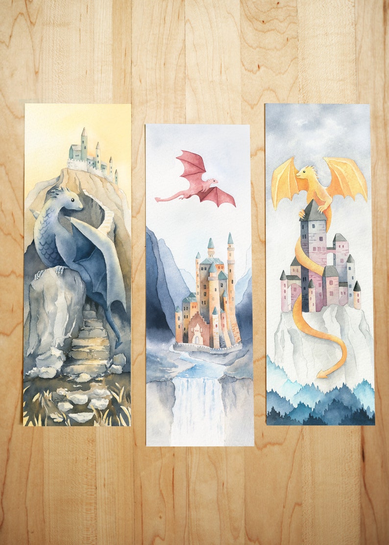 K&ouml;nnte beinhalten: Drei Aquarelle von Drachen und Burgen. Das erste Bild zeigt einen blauen Drachen, der auf einem Felsen sitzt, mit einer Burg im Hintergrund. Das zweite Bild zeigt einen rosa Drachen, der &uuml;ber einer Burg mit einem Wasserfall im Vordergrund fliegt. Das dritte Bild zeigt einen gelben Drachen, der sich um eine Burg mit einem Gebirge im Hintergrund schl&auml;ngelt.