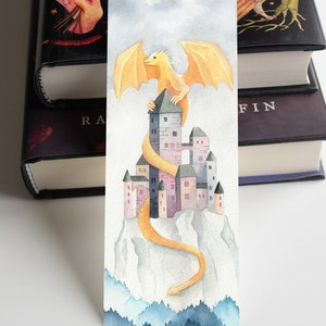 K&ouml;nnte beinhalten: Eine Aquarell-Illustration eines gelben Drachen, der auf einem rosa und grauen Schloss auf einem Berggipfel sitzt. Der Schwanz des Drachen schlingt sich um das Schloss und den Berghang hinunter. Der Hintergrund ist ein blauer und grauer Aquarellhimmel mit einem Wald am unteren Rand.