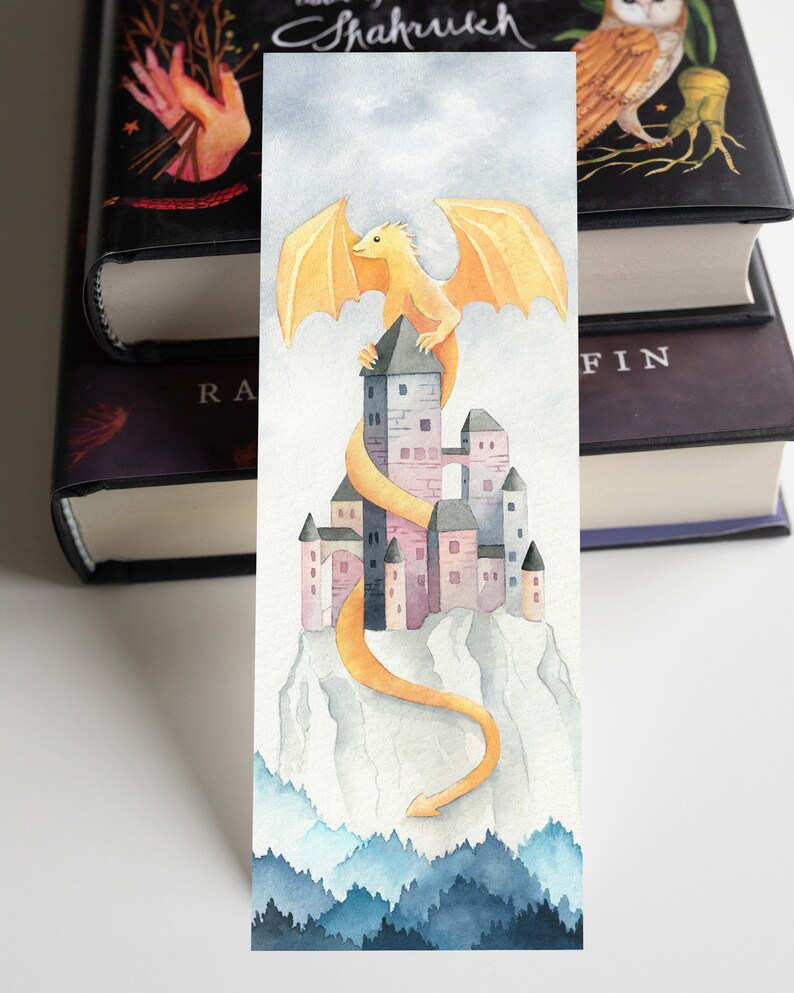 K&ouml;nnte beinhalten: Eine Aquarell-Illustration eines gelben Drachen, der auf einem rosa und grauen Schloss auf einem Berggipfel sitzt. Der Schwanz des Drachen schlingt sich um das Schloss und den Berghang hinunter. Der Hintergrund ist ein blauer und grauer Aquarellhimmel mit einem Wald am unteren Rand.