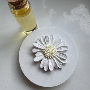 May include: White ceramic diffuser with a white daisy flower on top. A small glass bottle with a cork top filled with a yellow liquid is next to the diffuser.