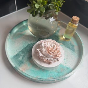 May include: A round, teal and white tray with a white and pink flower-shaped diffuser and a small glass bottle with a cork stopper.