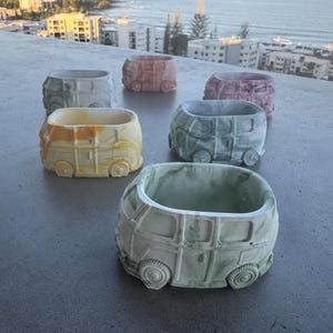 May include: A set of six colourful ceramic planters shaped like vintage Volkswagen vans. The planters are in various shades of blue, green, yellow, pink, and grey. Each planter has a unique marbled design.